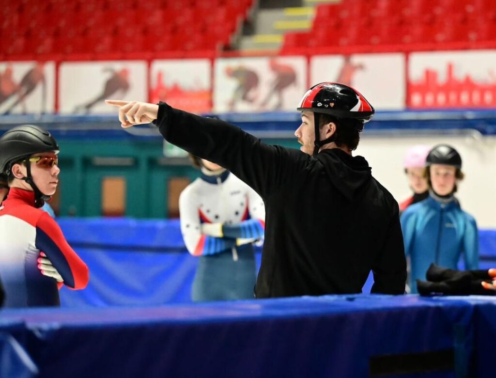 Samuel Gallant, casque sur la tête, pointe une direction à de jeunes patineurs de vitesse dans un aréna.